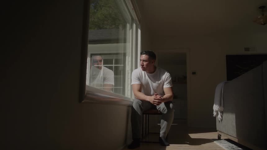 A young man sits contemplatively by the window in a living room during daytime