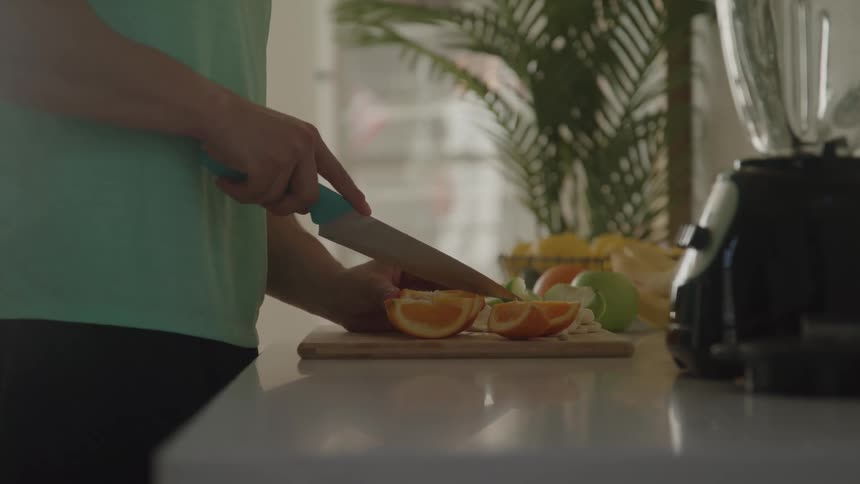 A person wearing a green shirt is standing at a kitchen counter and cutting an orange on a wooden cutting board with a knife while a blender sits next to them