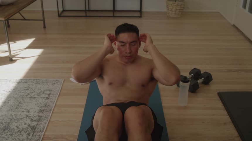 A man with no shirt on wearing black shorts is lying on a blue yoga mat on a wooden floor in a room doing situps while holding his hands behind his head