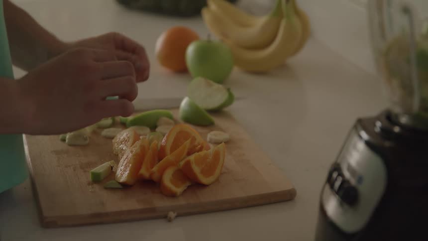A person wearing a green shirt is standing at a kitchen counter and cutting various fruits on a wooden cutting board and putting them into a blender