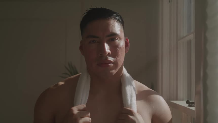 A shirtless young man inside a bathroom with a hand towel draped over his neck at midday