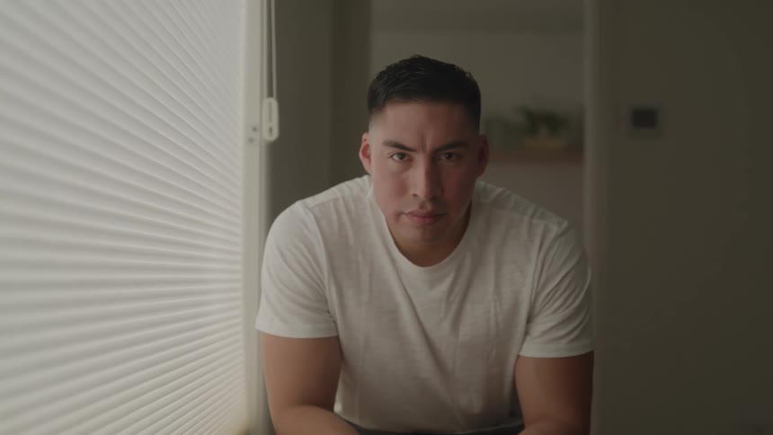 A man with an intense and serious expression sits in a bedroom looking directly at the camera