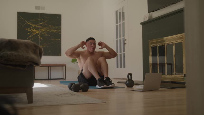 A man wearing trunks does situps shirtless in his living room