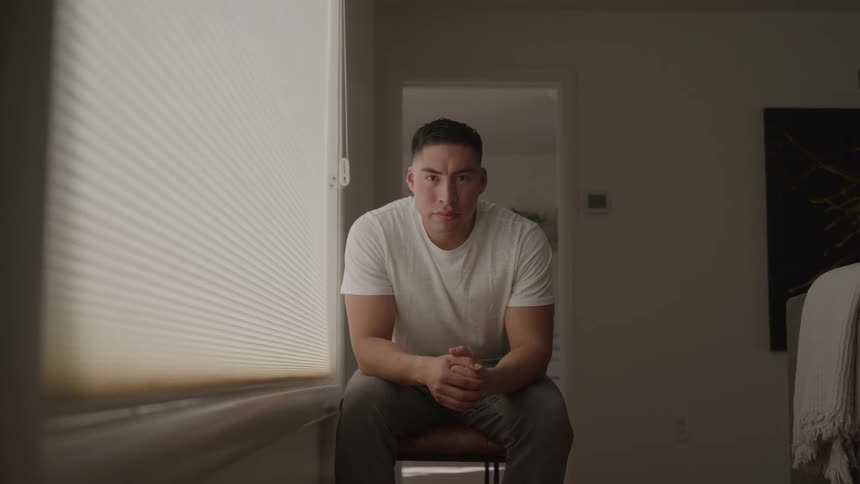 A man with a serious expression sits on a chair in a sunlit bedroom looking directly at the camera
