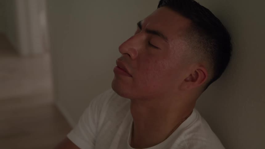 A close view shows a man with short dark hair wearing a white shirt as he leans against a wall and closes his eyes with a relaxed expression on his face