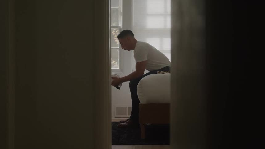 Man sitting on the edge of the bed putting on shoes in a room lit by the window