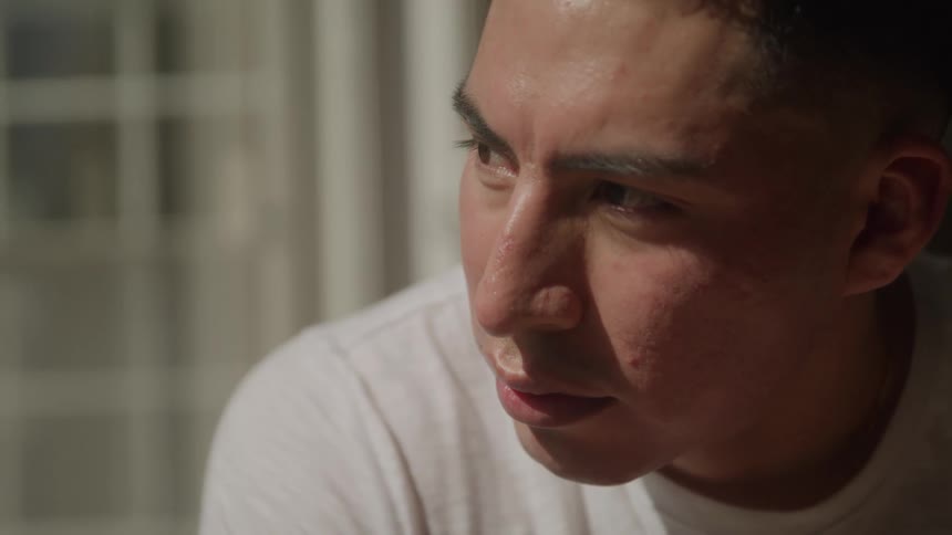 A person with a serious expression and a dark complexion looks downwards and to the side they are wearing a white tshirt and appear to be concentrating on something out of frame