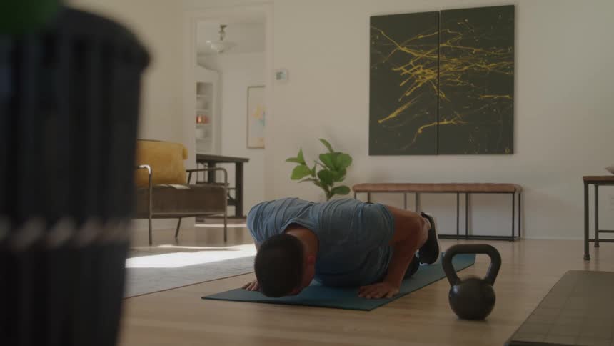 A man performs pushups on a blue exercise mat using dumbbells as handles in a room with a wooden floor and home furniture in the background