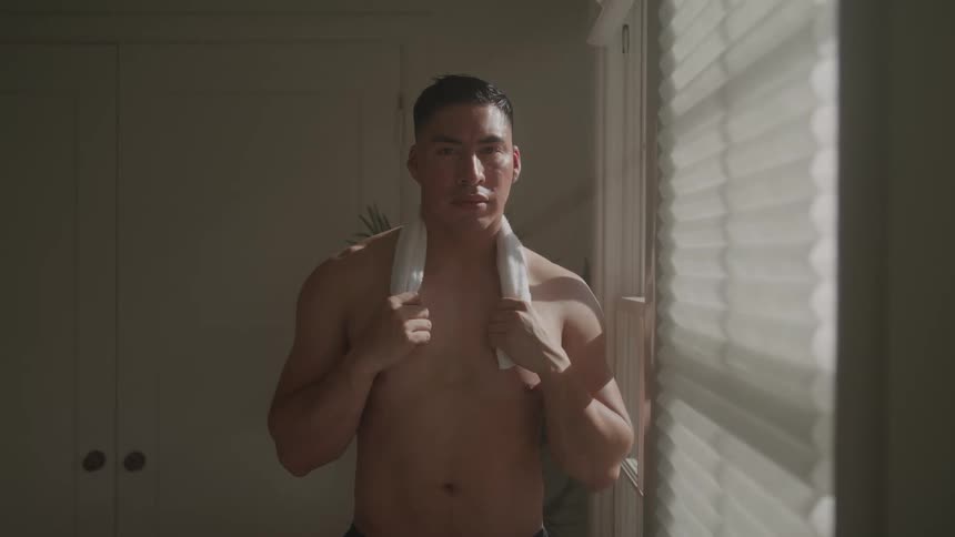 A shirtless young man stands indoors beside a window blind holding a face towel over his shoulder at midday