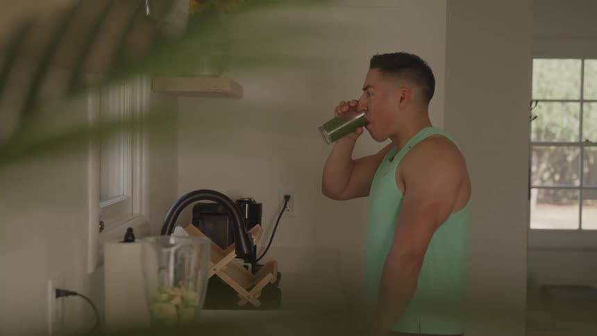 A man in a light green tank top stands in a kitchen holding a glass and drinking a green smoothie