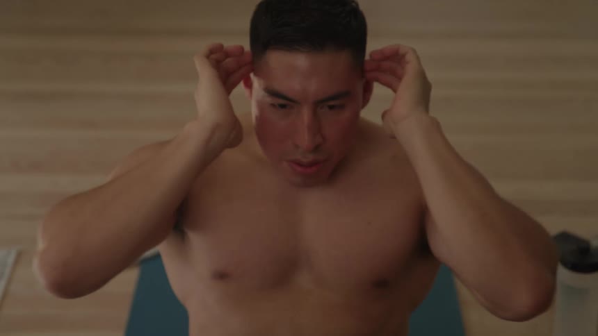 A shirtless man with his hands behind his head performs crunches on a blue exercise mat in a room with a wooden floor