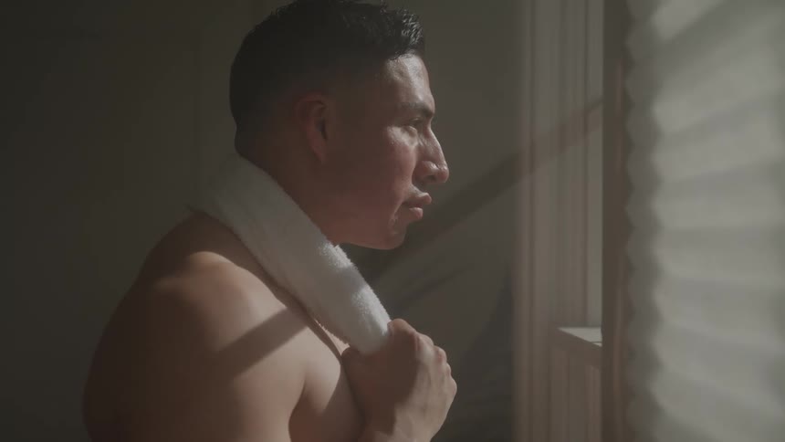 Shirtless man with a towel on the back of his neck looking out the window