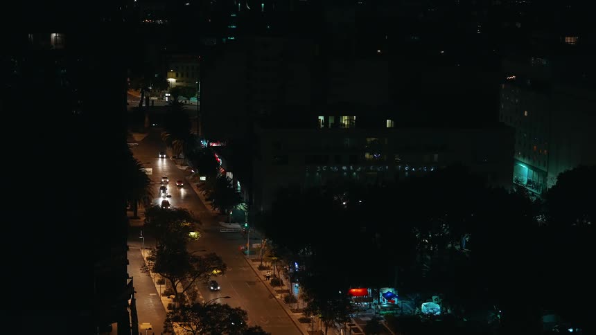 A topdown view shows cars driving along a street at night the buildings and trees are dark silhouettes against the city lights