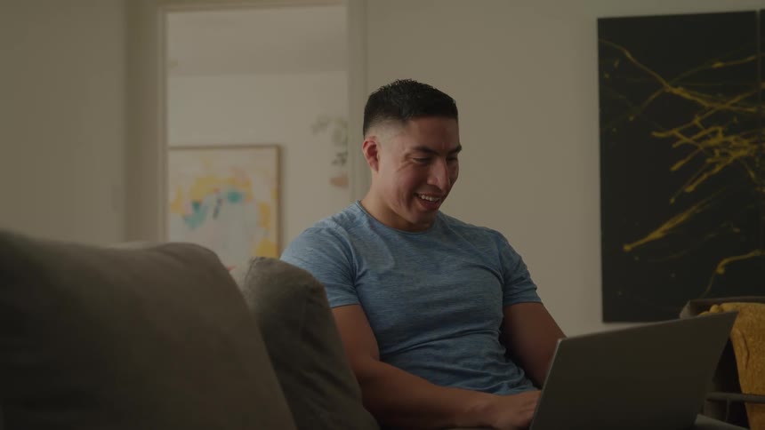 Young man with a blue tshirt sitting on a gray couch is sitting with his lap top he is typing and smiling as he stares at the lap top monitor there are paintings on the walls in the room with him and the room adjacent