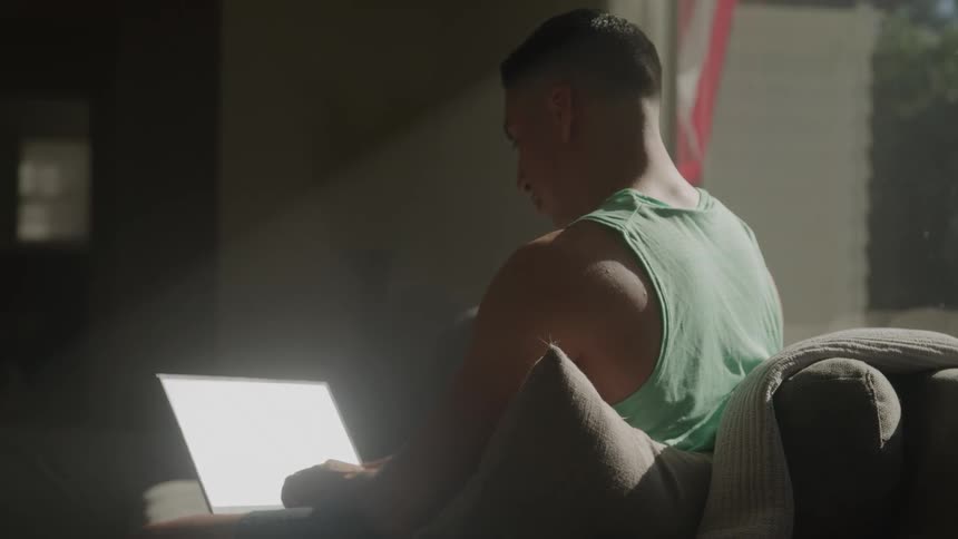A young man uses a laptop while sitting on a couch in a living room during daytime