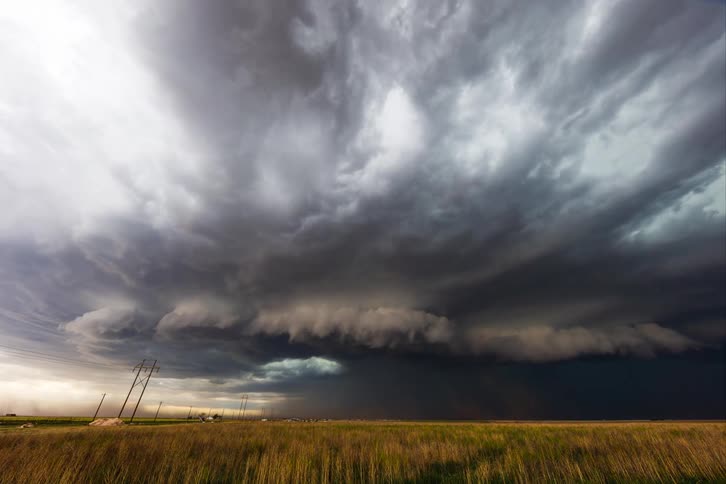 A massive dark storm cloud sweeps across a flat grassy plain with leaning utility poles hinting at powerful winds