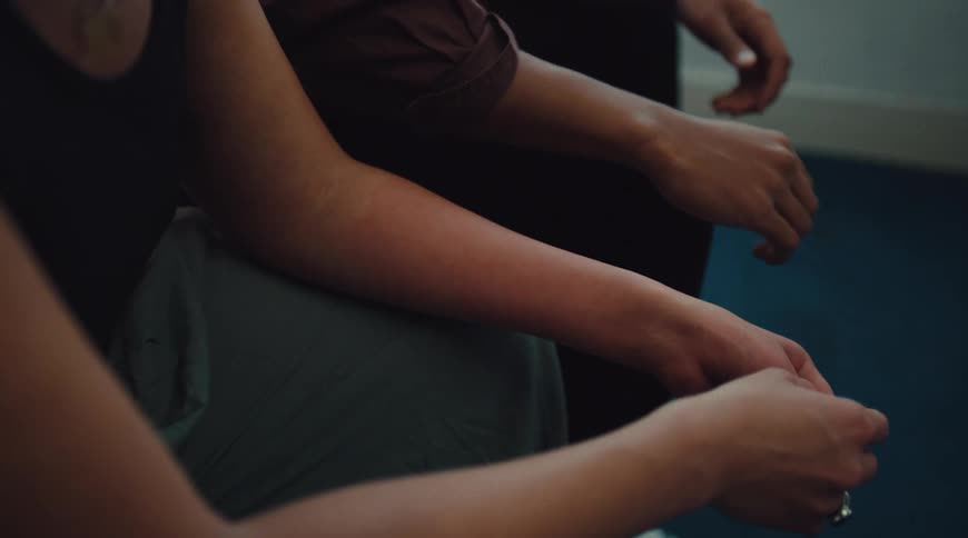 A man and woman sit side by side indoors as the woman reaches toward the man to hold his hand but stops halfway retreating her arm