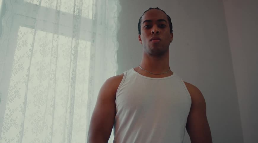 A young angry man with braided hair wearing an undershirt stands beside a window curtain at midday