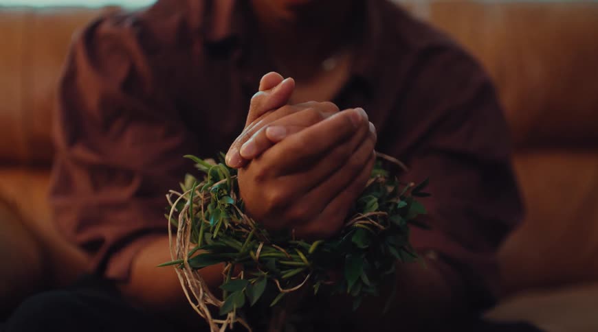 Person sits with clasped hands wrapped in green leaves and twine