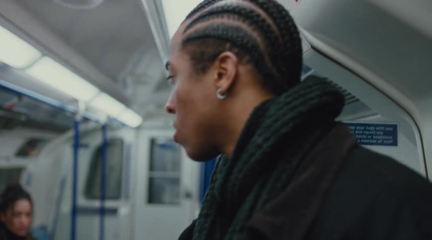 A young man with braided hair wearing a jacket and earrings stares at a woman sitting at a distance from him who is wearing a green coat during a train ride
