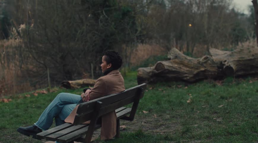 A young woman wearing a brown coat jeans and boots sits alone on a public park bench observing her surroundings at midday