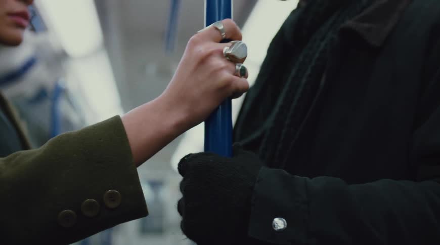 A woman wearing a coat and multiple rings on her right hand holds a pole inside a train with a young man in a jacket standing beside her during the ride