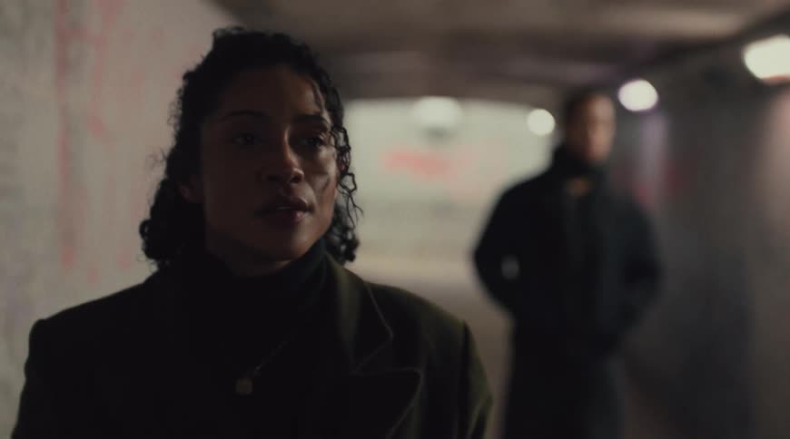 A man and a woman walk through a tunnel the woman wearing a coat and the man a jacket as she moves ahead while he walks slowly behind
