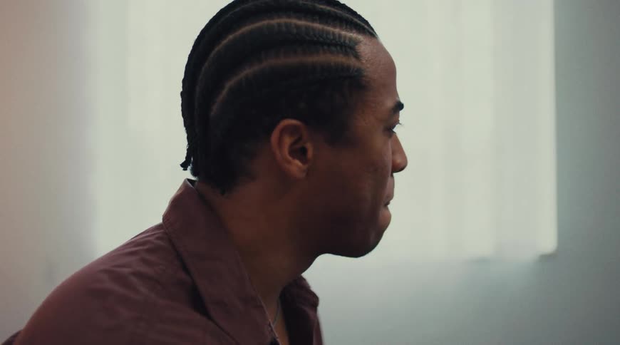 A young man with braids wearing a brown shirt sits indoors at midday looking to the right suspiciously
