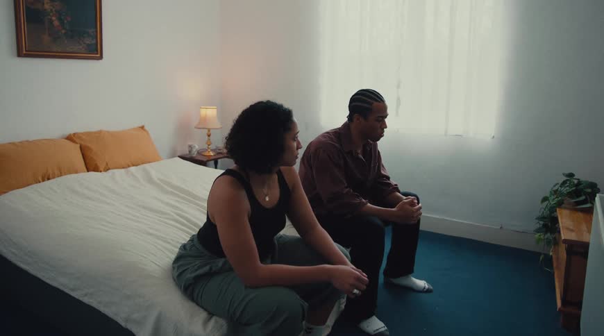 A man and a woman sit in a bedroom initially facing forward then turning to look at each other as if in conversation