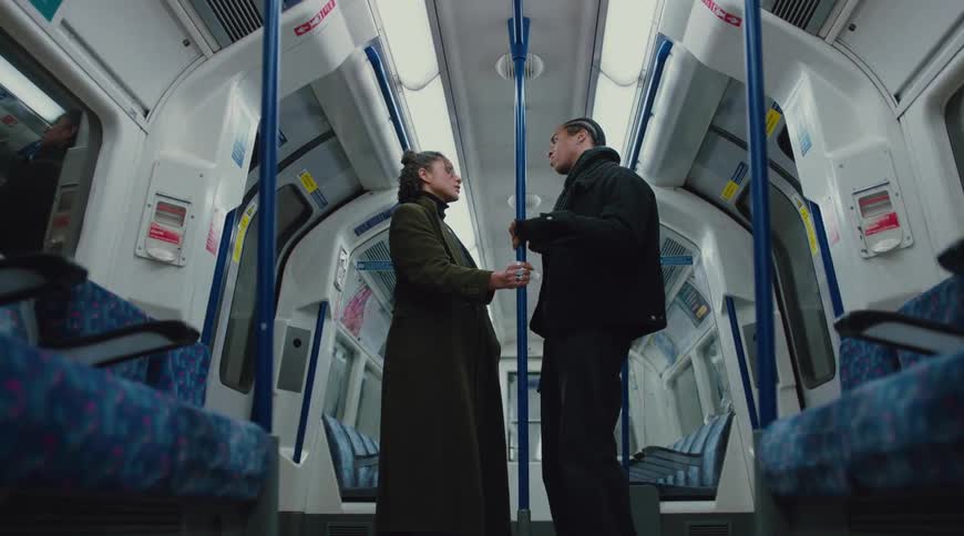 A young man with braided hair wearing a jacket and a woman in a green coat stand inside a moving train car walking toward each other while holding the poles and looking at each other