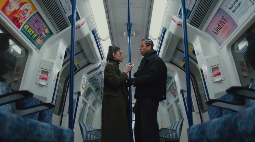 A young woman with curly hair wearing a green overcoat and a necklace walks toward a man with braided hair holding a pole in a train car during a ride speaking to him and looking sad afterward