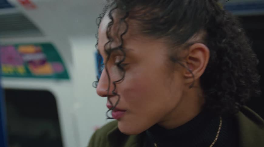A young woman with curly hair looks up then down and then to the side wearing a green coat and a black turtleneck with a gold necklace
