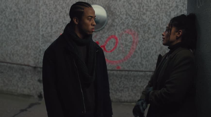 A young woman wearing a coat and a man with braided hair wearing a jacket walk down the stairs into an underground subway stopping to have a discussion midday