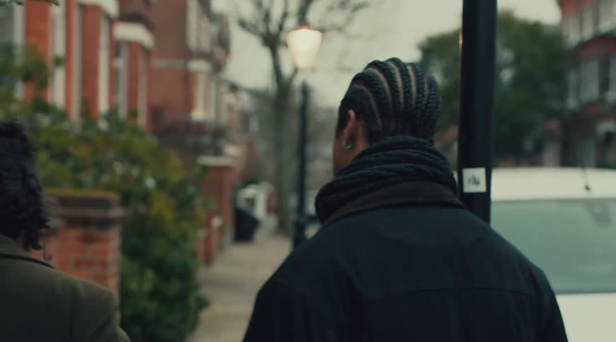 A young man walks beside a young woman on a residential city sidewalk both wearing winter jackets as he tries to catch up and talk to her while she ignores him