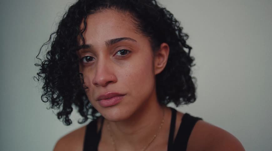 A young woman with curly hair sits indoors looking slightly sad