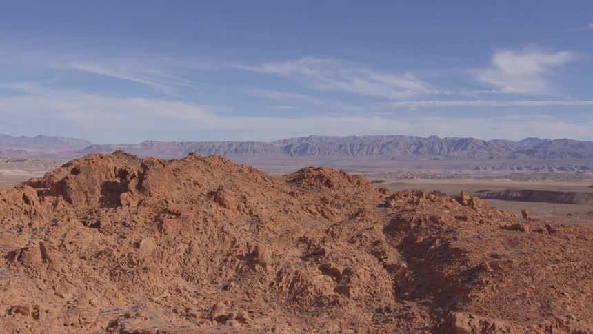A panoramic shot captures a vast arid landscape under a clear blue sky with sparse clouds in the foreground a prominent redbrown rock formation dominates the view with distant mountain ranges visible in the background