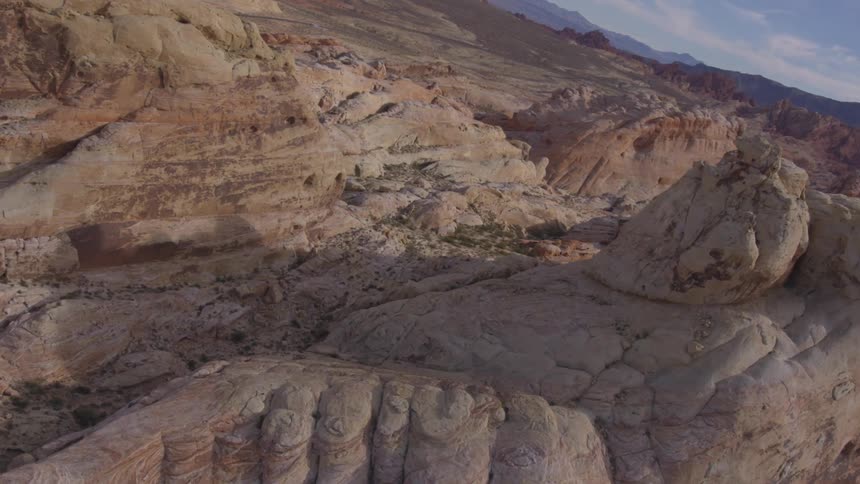 An aerial view shows a vast rocky desert landscape with varying shades of red orange and white under a clear sky with scattered clouds