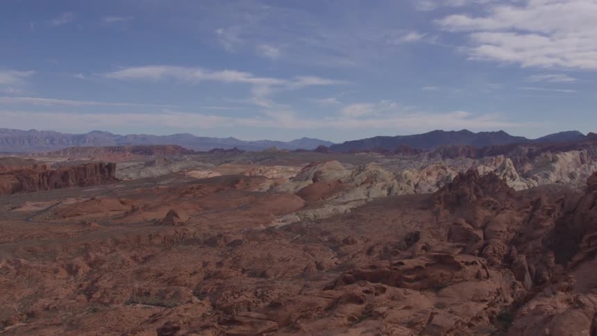 An expansive aerial shot shows a rugged desert landscape with dramatic mountain ranges under a partly cloudy blue sky the camera pans slowly across the rocky terrain highlighting the striking contrast between the redbrown formations in the foreground