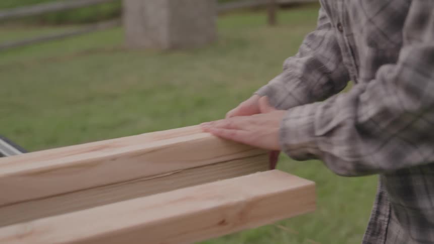A medium shot shows a persons torso and arms as they carefully handle two long wooden planks the person is wearing a gray plaid shirt and is standing in a grassy area with the planks resting on what appears to be the back of a truck