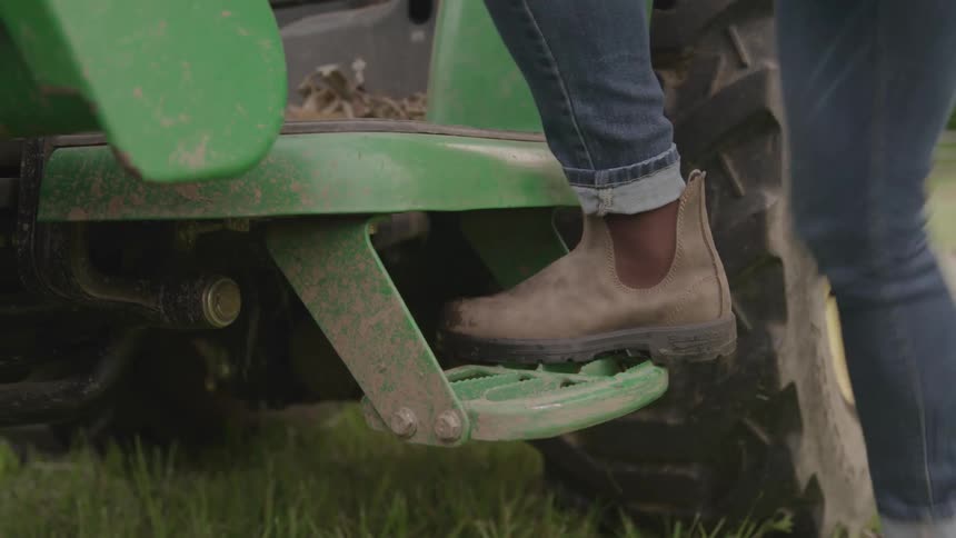 A lowangle shot focuses on the weathered step and tire of a green tractor parked on a grassy field a person wearing blue jeans and brown boots steps onto the tractor planting their foot firmly on the metal step