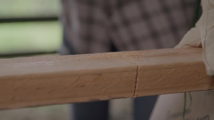 A gloved hand uses a handsaw to cut through a piece of lightcolored wood