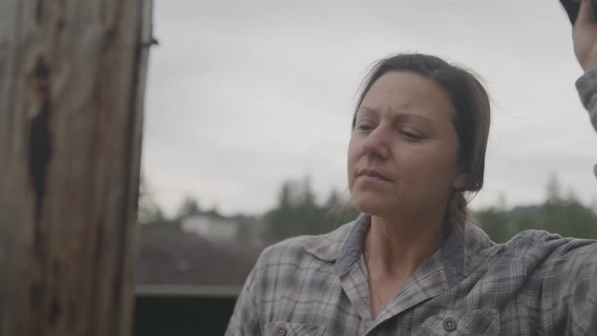 A woman in a green cap and plaid shirt hammers a nail into a wooden post outdoors