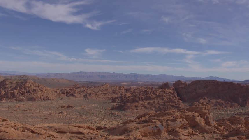 A vast desert landscape featuring numerous red and orange rock formations in the foreground and a hazy mountain range under a blue cloudy sky