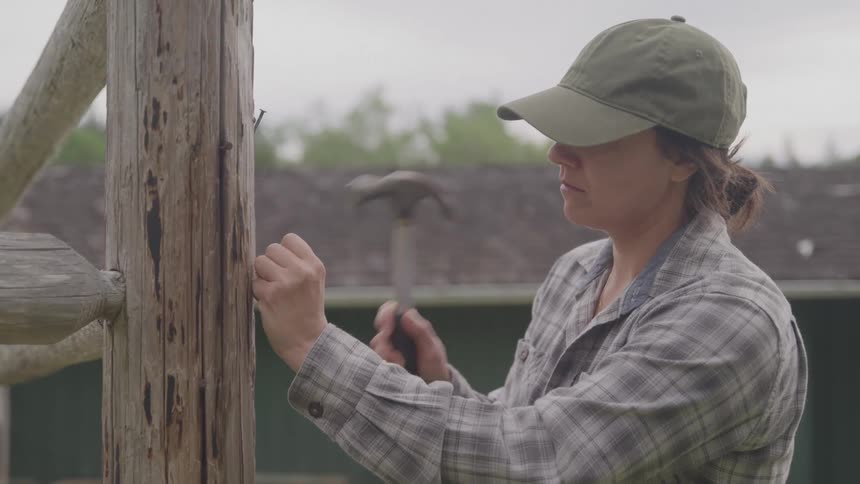 A woman in a green cap and plaid shirt hammers a nail into a wooden fence post outdoors