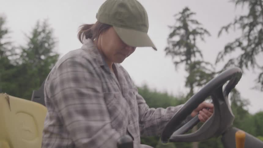 A medium shot captures a woman sitting in the drivers seat of a green tractor holding the steering wheel she is dressed in a plaid shirt and a baseball cap and she reaches for a lever on the dashboard appearing to get ready to operate the vehicle