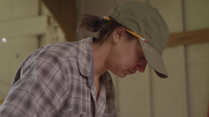A video shows a closeup of a woman wearing a baseball cap and a plaid shirt looking down intently she appears to be concentrating on a task with a pencil tucked behind her ear