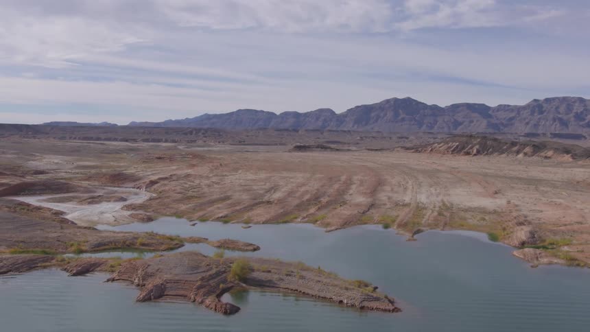 A wide view of a rocky desert landscape with sparse vegetation bordered by calm blue water and distant mountains under a partly cloudy sky