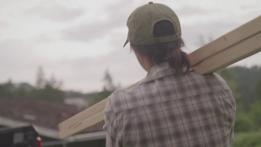 A woman in a plaid shirt carries a long wooden beam on their shoulder outdoors to place in a truck