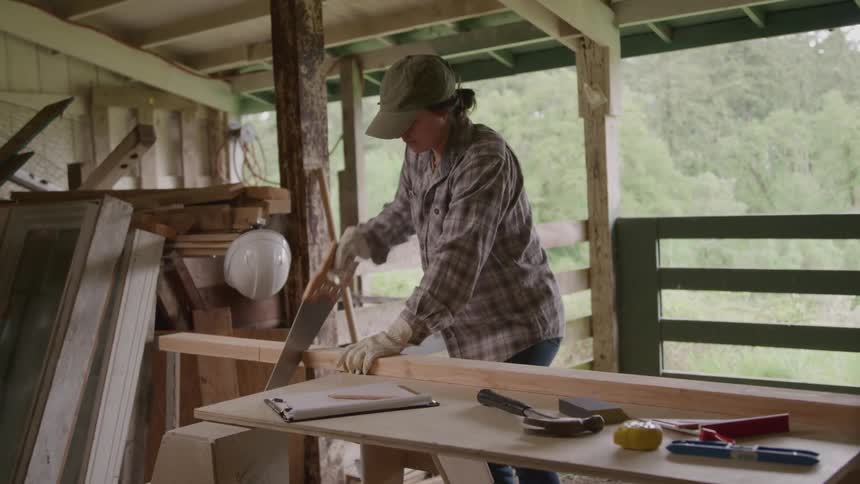 A person in a plaid shirt and a baseball cap is seen working in a rustic barn or workshop they are using a handsaw to cut a piece of lumber on a wooden workbench with various tools and materials scattered around them