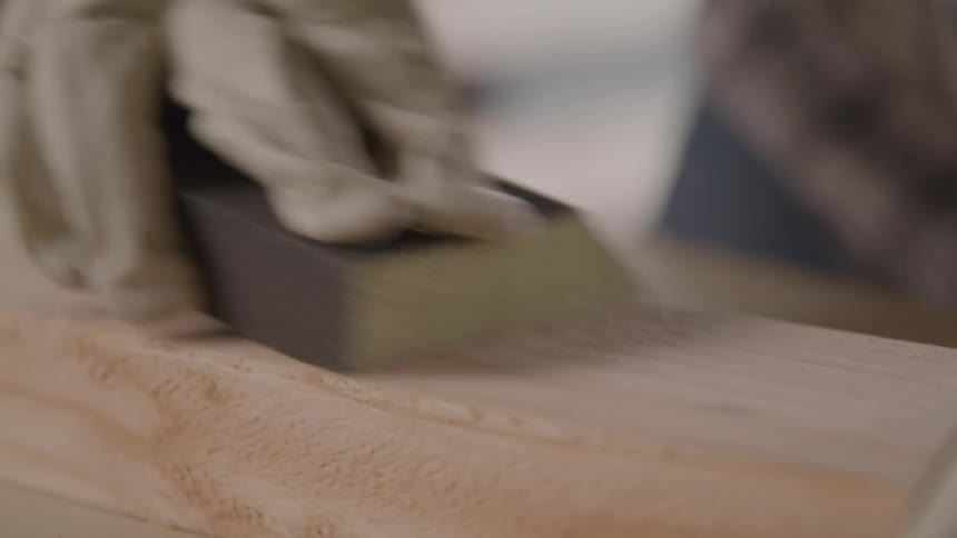 A gloved hand sands the surface of a wooden board for a smooth finish